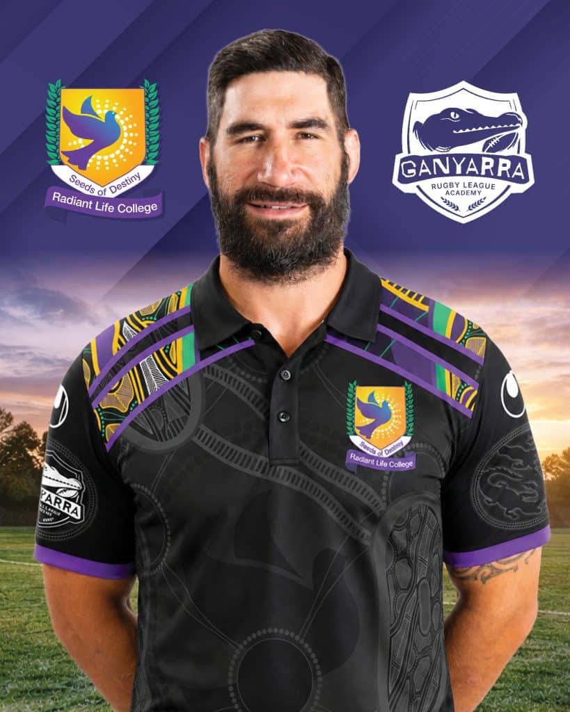 Man wearing black shirt with school logos on left chest and shoulder stripes standing between two sports logos against a purple sky backdrop