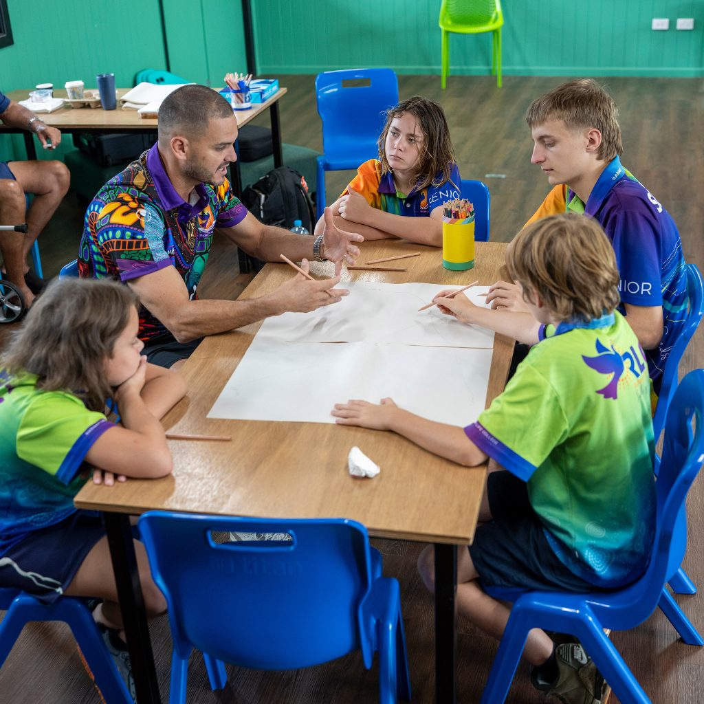 Group of children and an adult seated at a wooden table drawing with pencils in a bright classroom with blue chairs