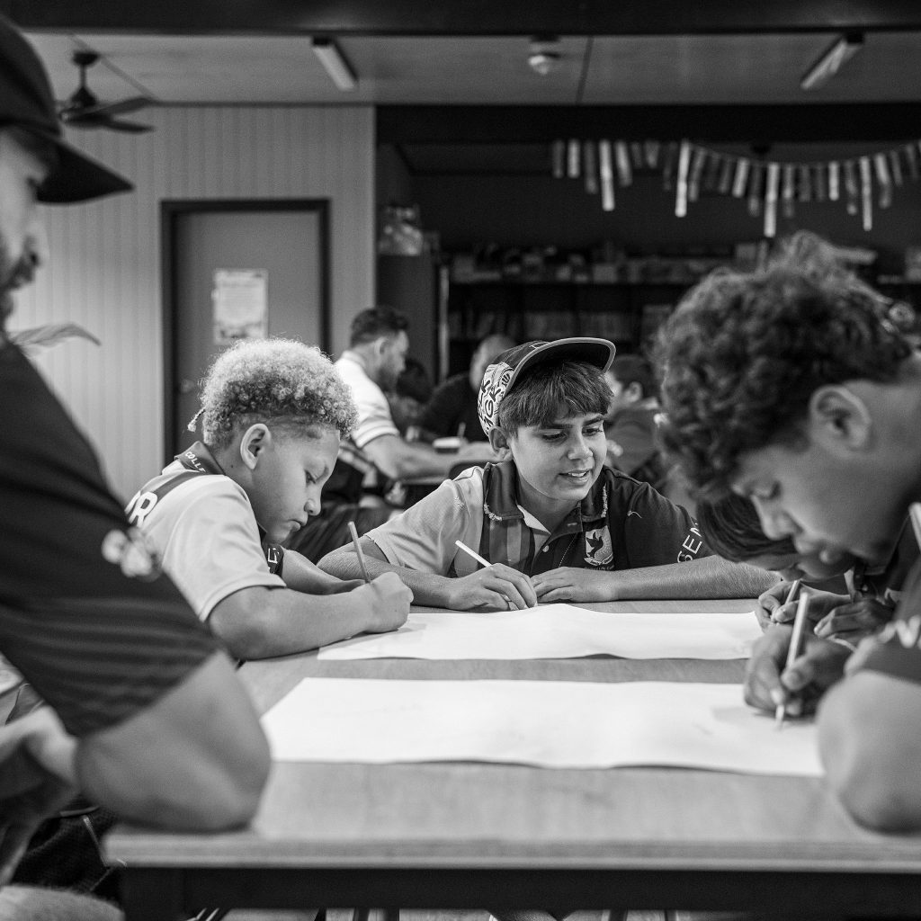 People gathered around a table drawing or writing on large sheets focusing on their paper work in a workshop setting black and white