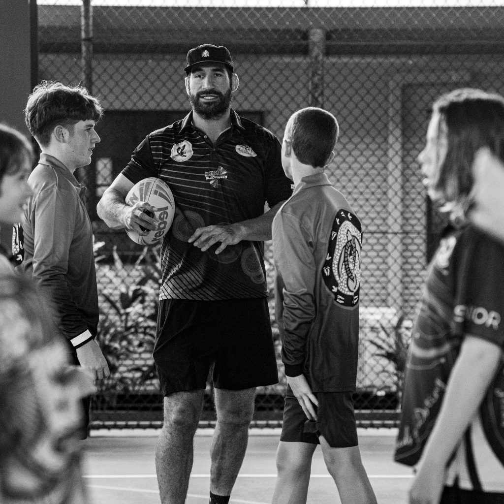 Coach in a striped shirt and cap speaks to players on an indoor court holding a rugby ball in front of a chain link fence