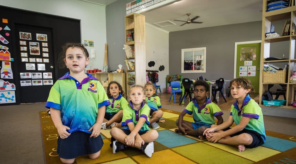 Group of young students in green uniforms sitting on a colorful classroom rug indoors facing the camera