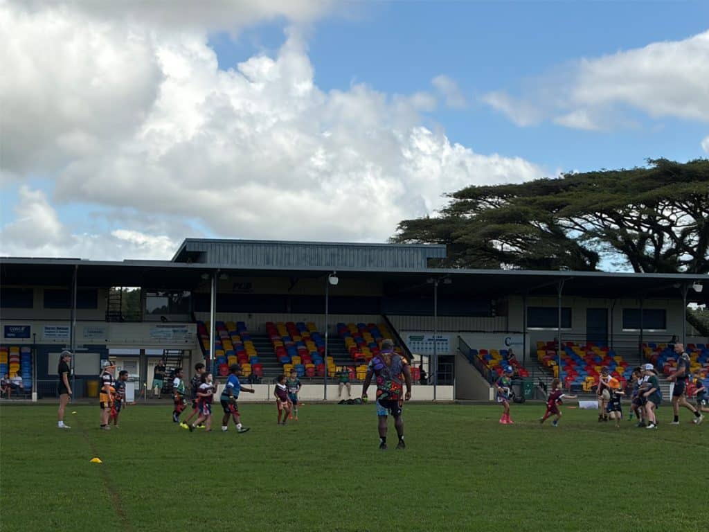 Kids playing rugby on a grassy field with a stadium and colorful empty seats in the background under a cloudy sky